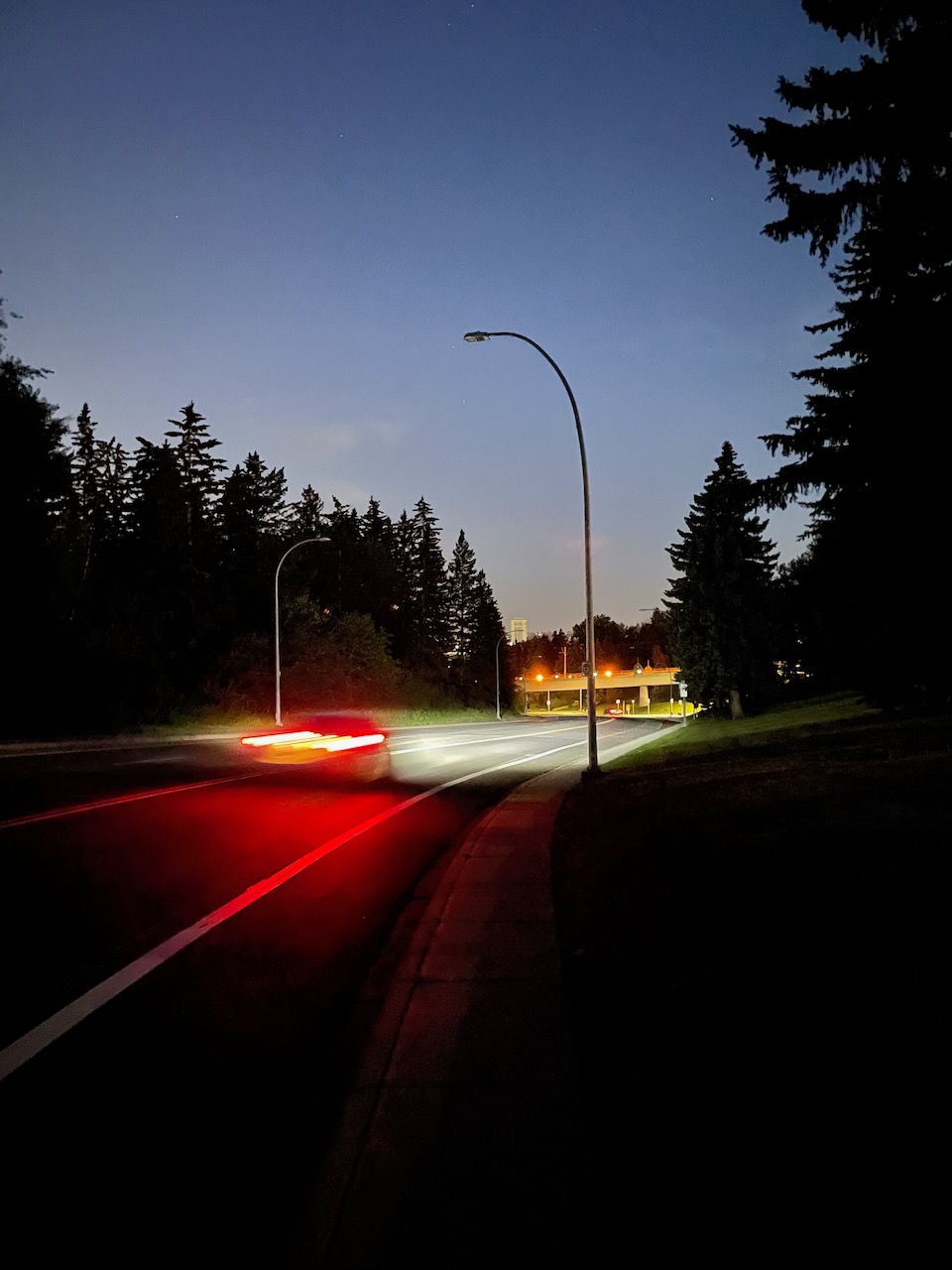 Photo at night of a dark hilly street and a taillight streak from a passing car.