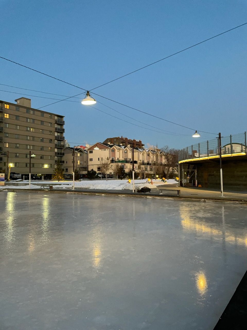Photo at dusk of an empty outdoor ice rink with overhead industrial-style lights on cables.